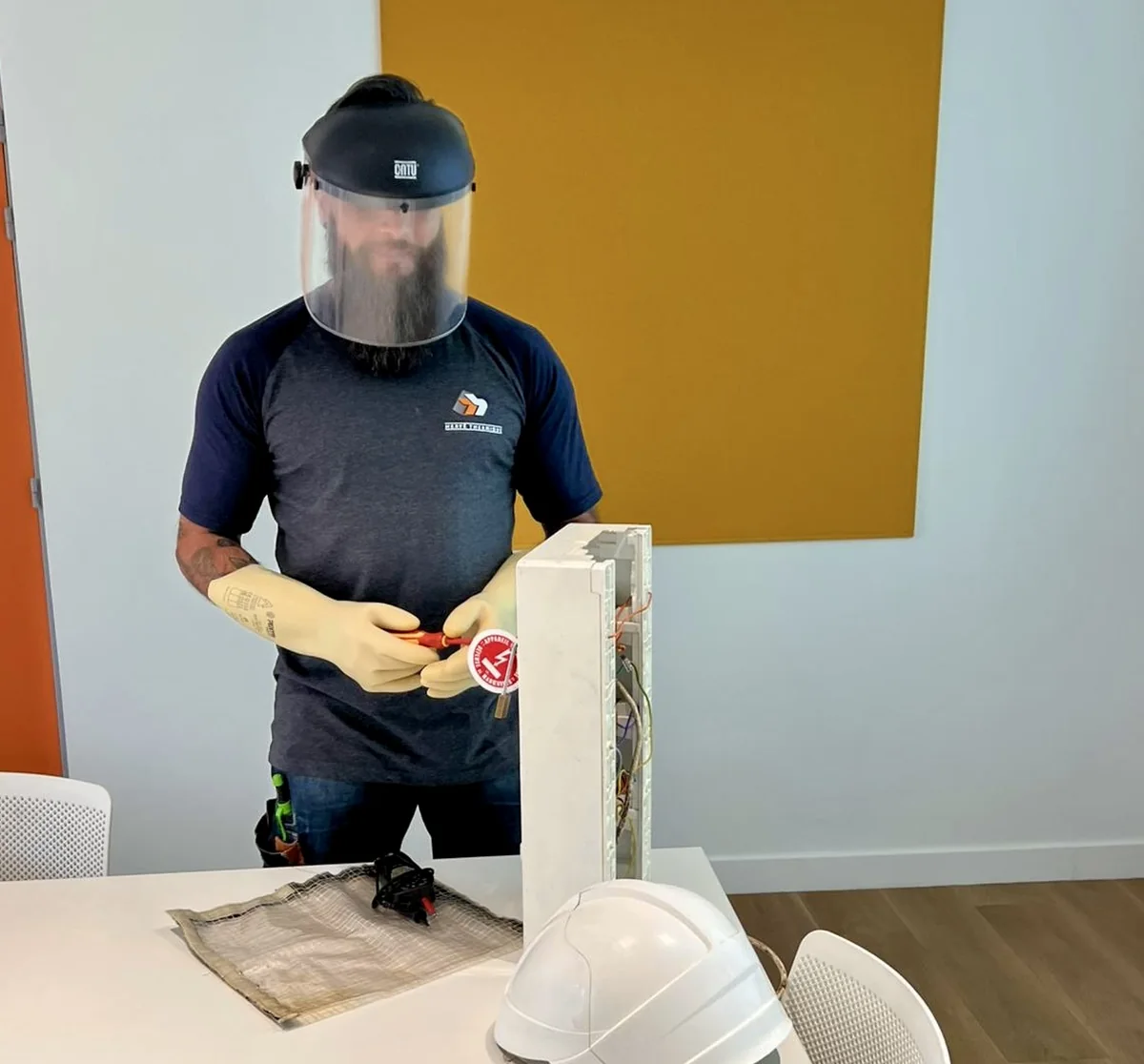 Stagiaire équipé d'un écran facial et de gants isolants intervenant sur un tableau électrique pédagogique, avec tapis isolant et vérificateur d'absence de tension, formation habilitation électrique NB Formation